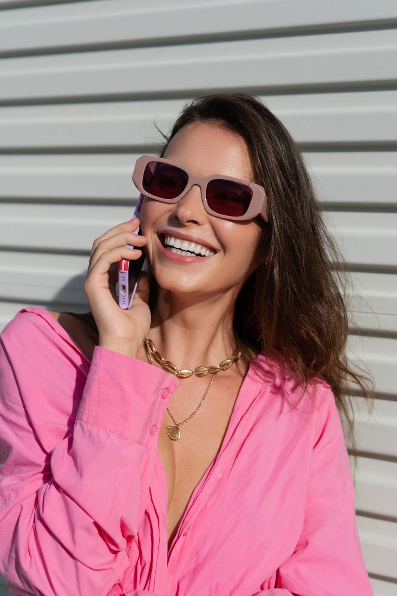 Young beautiful brunette woman in a pink shirt, neck jewelry, necklace, trendy sunglasses on the background of a light garage fence, calls the phone, laughs, smiles